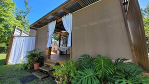 Um pequeno edifício térreo com telhado de metal corrugado e varanda coberta. Escadas levam à varanda, e a área é cercada por vegetação exuberante, incluindo plantas tropicais grandes.