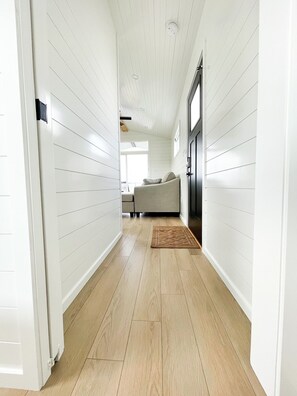 A hallway with white paneled walls, a wooden floor, and a sofa in the distance.