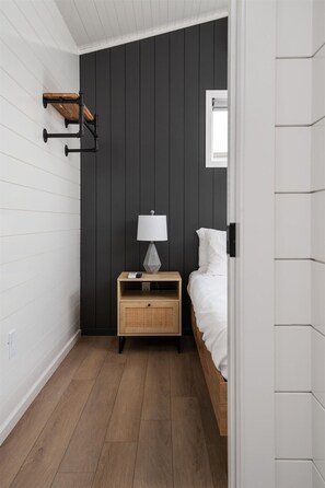 A small, modern bedroom with a dark wall, a wooden nightstand, a white lamp, and a bed with white bedding.