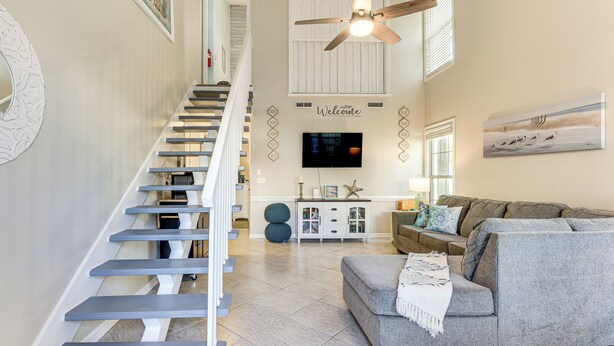 A modern living room with a grey sofa, a staircase with white railings, a ceiling fan, and a flat-screen TV mounted on the wall. - Walk to Beach! PCB Resort Condo w/ Pool Access (Panama City Beach)