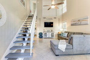 A modern living room with a grey sofa, a staircase with white railings, a ceiling fan, and a flat-screen TV mounted on the wall.