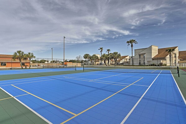 An outdoor tennis court with a blue playing surface and surrounding greenery, including palm trees and a building in the background.