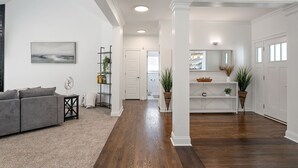 Un salon spacieux avec un canapé gris, une petite table d’appoint et une bibliothèque. Un couloir mène à une porte et à une salle de bain.
