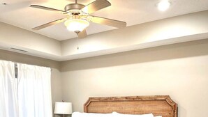 A bedroom with a wooden bed, white bedding, a colorful pillow, a ceiling fan, a white lamp, and a red rug.