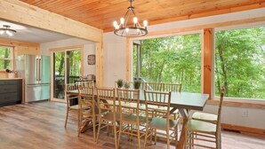 Una cocina amplia con armarios de madera y una isla grande, un comedor con sillas de bambú y una mesa, y ventanas grandes que ofrecen vistas al bosque.
