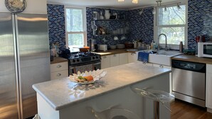 A modern kitchen with a stainless steel refrigerator, a gas stove, and a microwave. There is a window providing natural light, and the backsplash is made of blue tiles. - Catherine’s Place (Southampton)