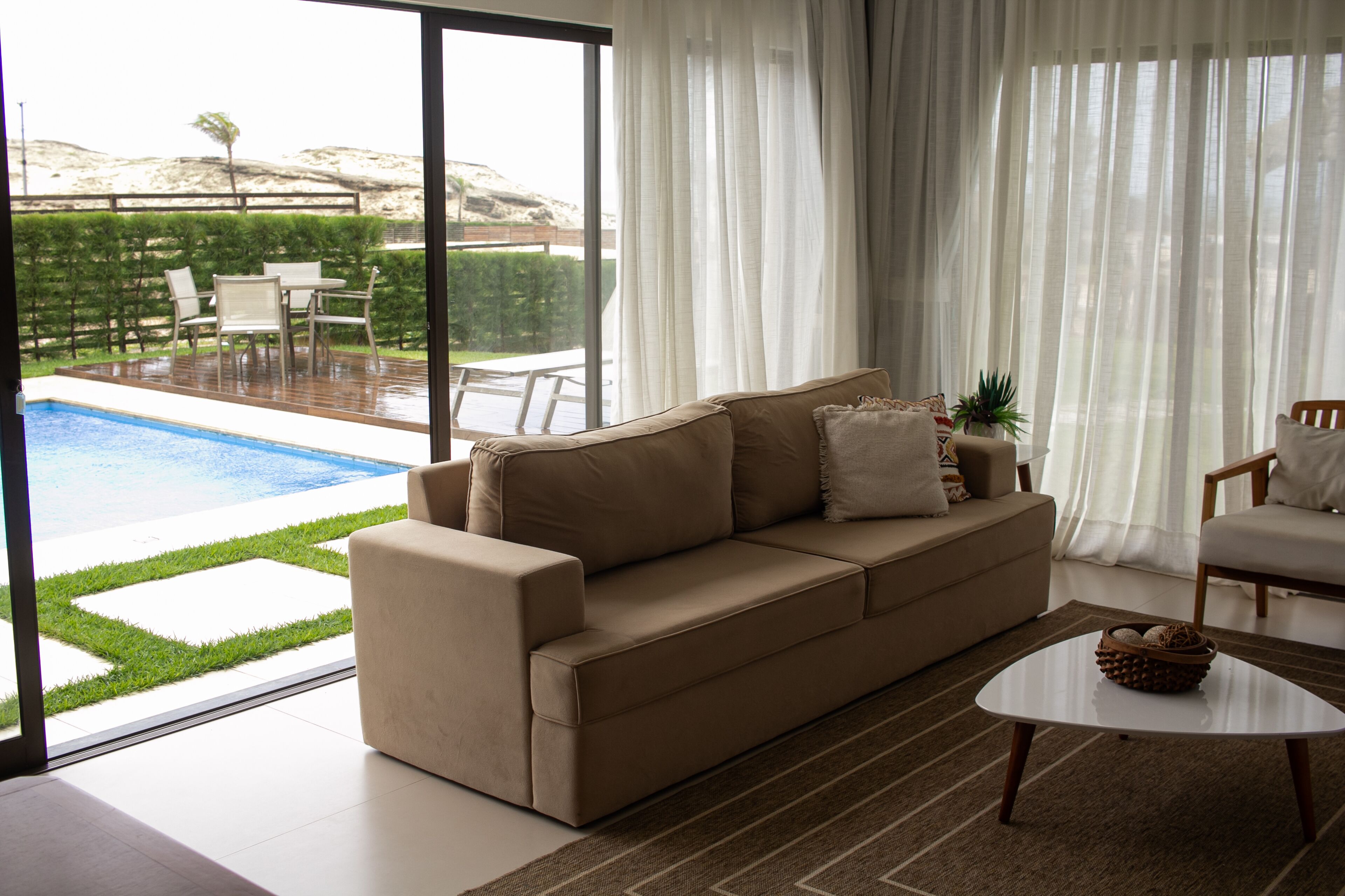 Un saló modern amb un sofà beix, una taula de centre i vista a una piscina i zona d'estar a l'aire lliure.