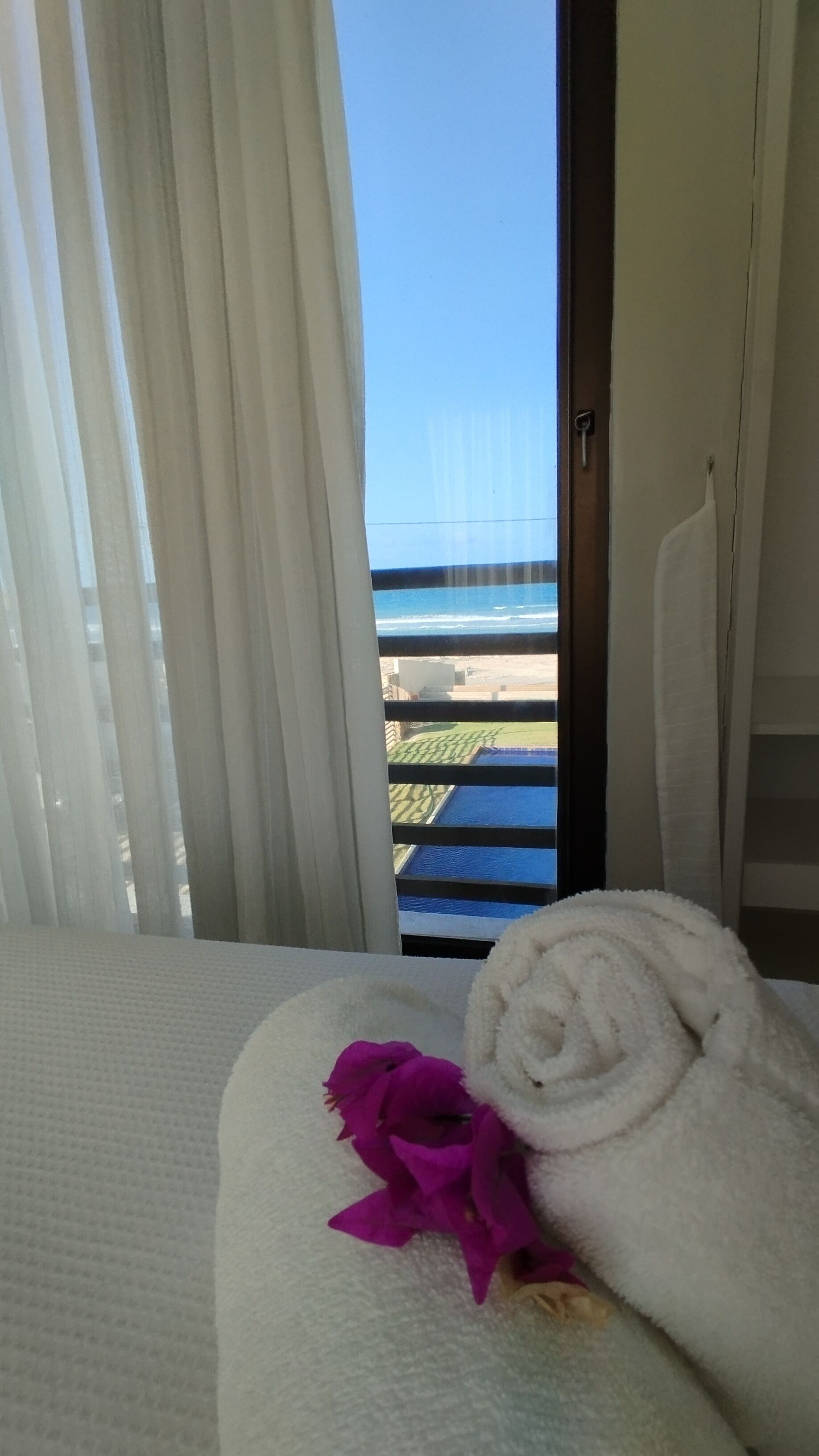 A hotel room with a view of the ocean through a window with white curtains.