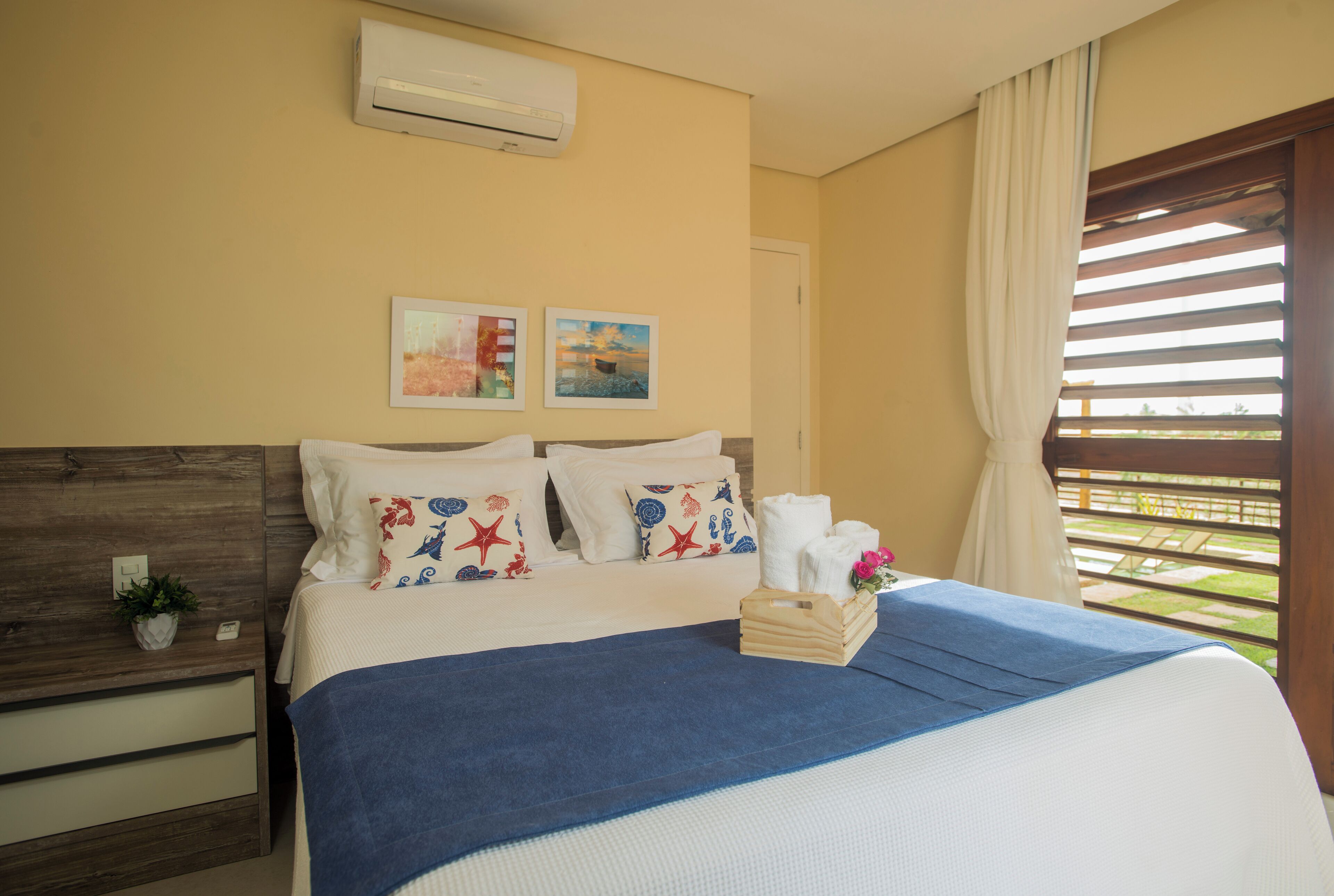 A hotel room with a bed, a nightstand, a potted plant, and a window with blinds.