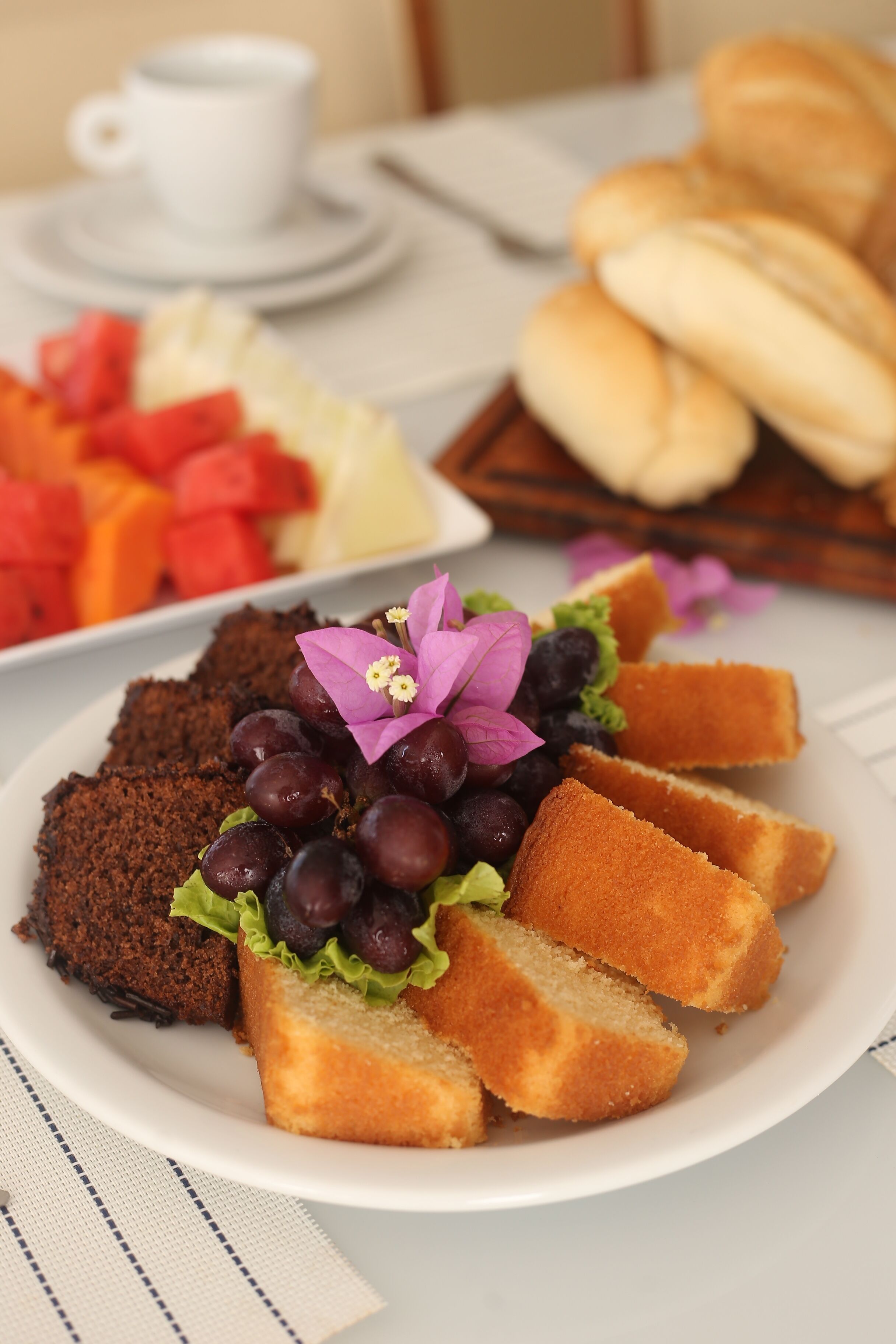A plate of food with bread, grapes, and a dark cake, along with a cup and a tray of bread rolls.