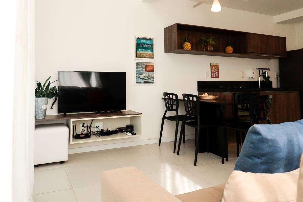 A modern living room with a flat-screen TV, a sofa, a dining area with chairs, and a kitchen with wooden cabinets.