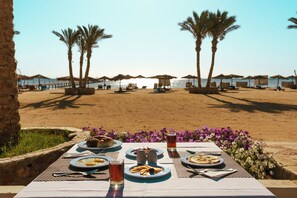 Ein gedeckter Tisch mit Speisen, Besteck und Gläsern, mit Blick auf einen Strand mit Palmen und Sonnenschirmen.