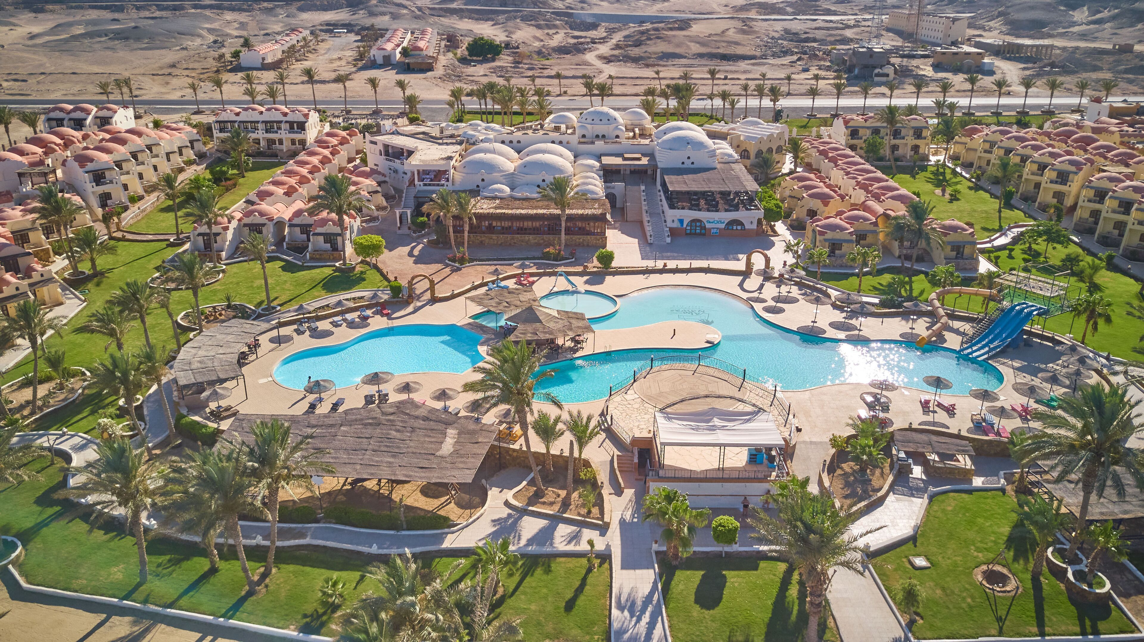 Vista aérea de um resort com várias piscinas, palmeiras e paisagem desértica ao fundo.