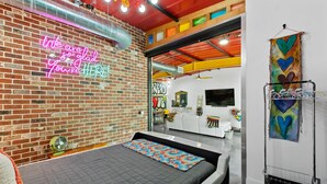 A modern hotel room with a brick wall, a neon sign, a bed with a patterned bedspread, and a colorful artwork on the wall. - The ArtHouse: Where Industrial Charm Meets Creative Inspiration (St. Augustine)