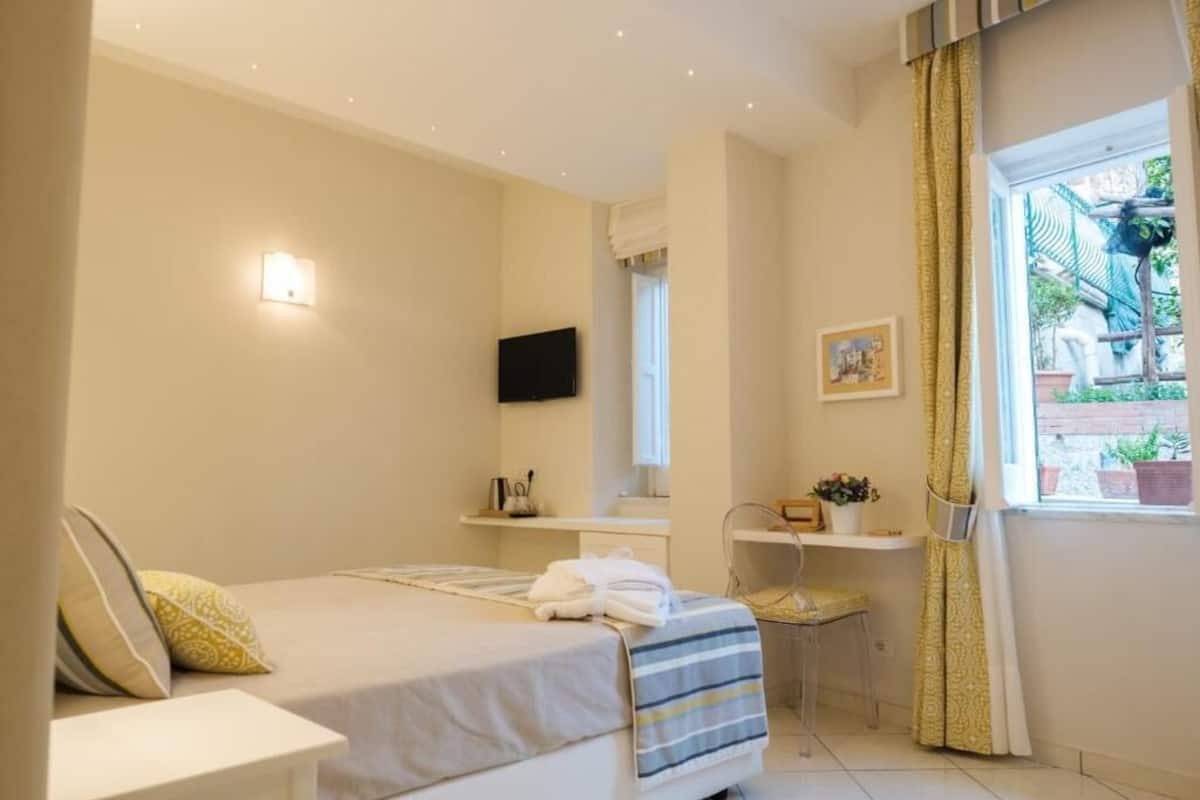 A bedroom with a bed, bedside table, a small desk, a chair, a TV mounted on the wall, and a window with curtains.