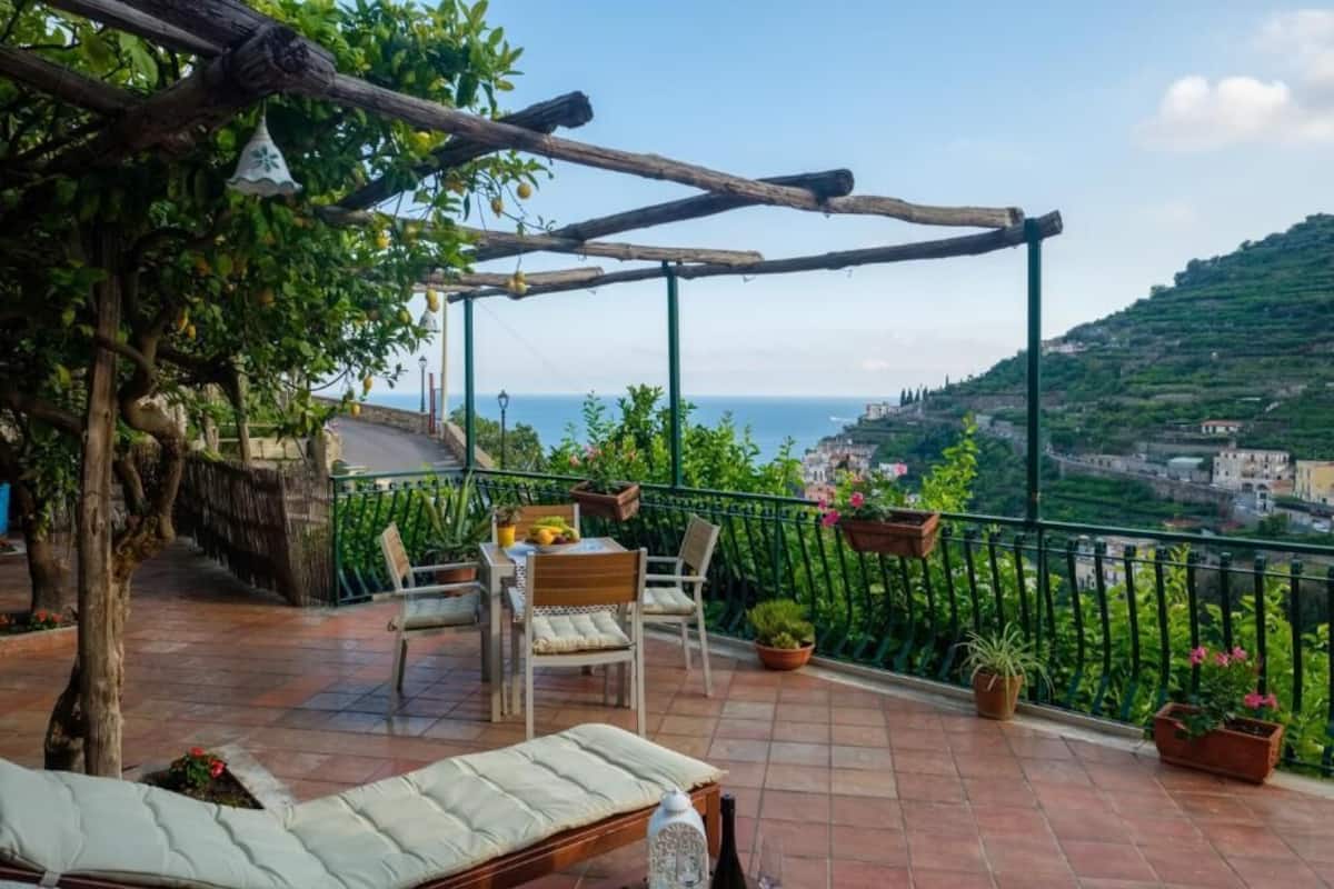 A terrace with a table and chairs, potted plants, and a view of the sea and hills.