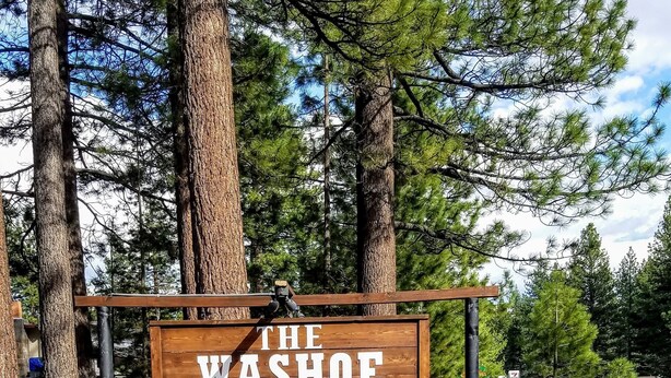 En träskylt med texten ”THE WASHOE 75 1 EMERALD BAY ROAD”, omgiven av träd och en bänk.