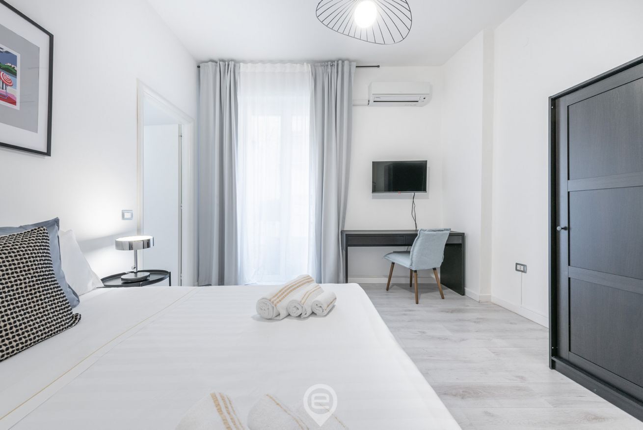 A modern hotel room with a large bed, a desk, a chair, and a TV.