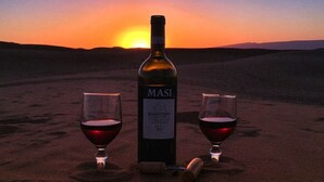 Een fles wijn en twee glaasjes op een zandig oppervlak, met een zonsondergang op de achtergrond.