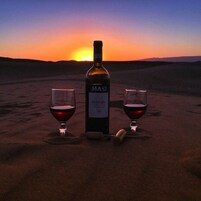 Una botella de vino y dos copas sobre arena, con un atardecer de fondo.
