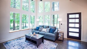 Ein geräumiges Wohnzimmer mit grauem Sofa, einem Couchtisch und einem großen Fenster mit Blick auf einen Wald.