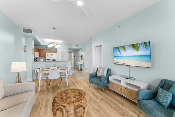 A modern living room with a dining area, kitchen, and a large TV displaying a beach scene.