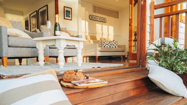 A cozy living room with a wooden floor, a dining table with a bottle of champagne, and a sign that reads "BEACH HOUSE". - Hamptons on Queen - Sunset Water Views (Bongaree)