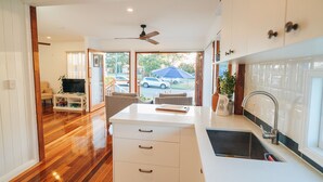 A modern kitchen with white cabinets, a built-in oven, and a sink. There is a dining area with a table and chairs, and a television in the corner. - Hamptons on Queen - Sunset Water Views (Bongaree)