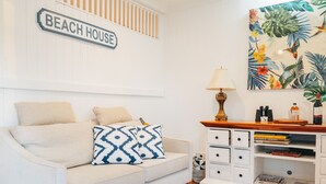 A living room with a beige sofa, patterned pillows, a white side table with a lamp, a bookshelf, and a wall sign reading "BEACH HOUSE". - Hamptons on Queen - Sunset Water Views (Bongaree)