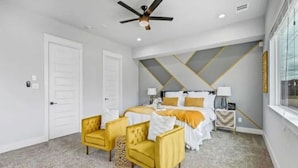 A bedroom with a bed, two yellow armchairs, a ceiling fan, and a nightstand with a lamp.