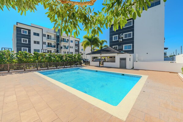 Una piscina con bordeado de baldosas, rodeada de edificios modernos y una palmera.