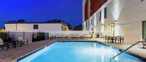 Una piscina interior con borde de azulejos, rodeada de sillas y mesas de descanso, junto a un edificio de fachada roja.