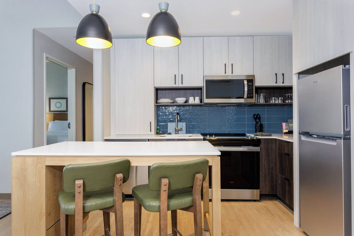 Una cocina moderna con una isla central, dos lámparas colgantes, un microondas, un refrigerador y sillas de madera.