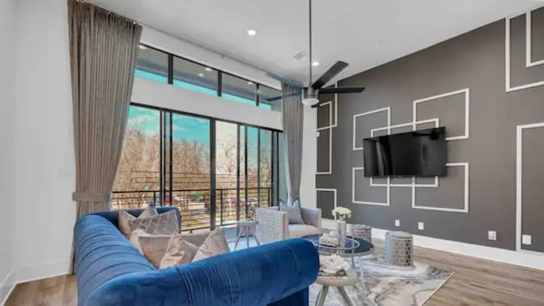 A modern living room with a blue sofa, a flat-screen TV mounted on the wall, and a large window offering a view of the outdoors. - Stylish Cozy home near DT Houston and Medical Cntr (Houston)
