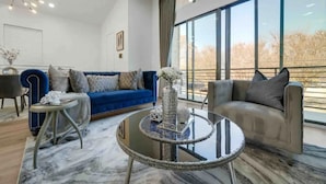 A modern living room with a blue sofa, a beige sofa, and a glass coffee table. There is a large window with a view of trees. - Stylish Cozy home near DT Houston and Medical Cntr (Houston)
