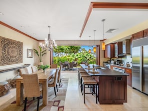 A modern kitchen with a large island, dining area with wicker chairs, and a view of lush greenery.