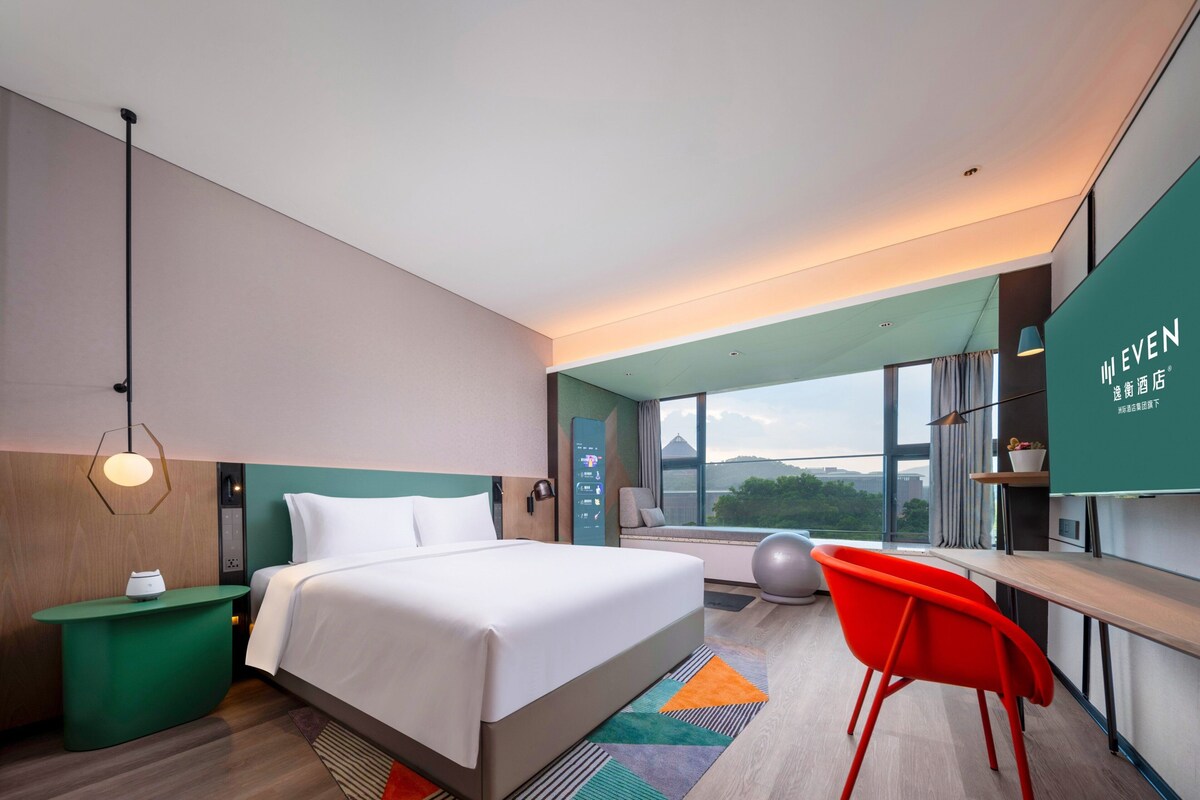 Una habitación de hotel moderna con una cama grande, un escritorio, una silla roja y vistas a la ciudad.