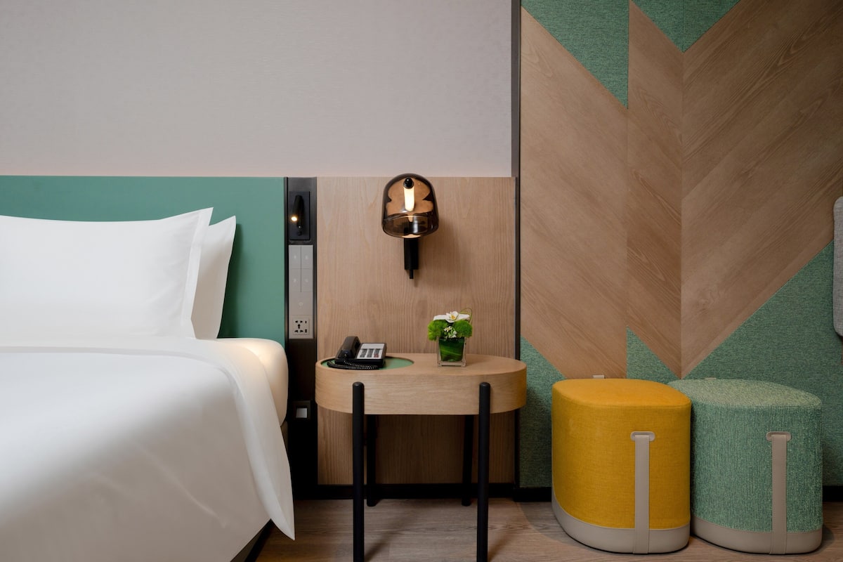 A hotel room with a bed, a small table with a phone and a vase, and two ottomans.