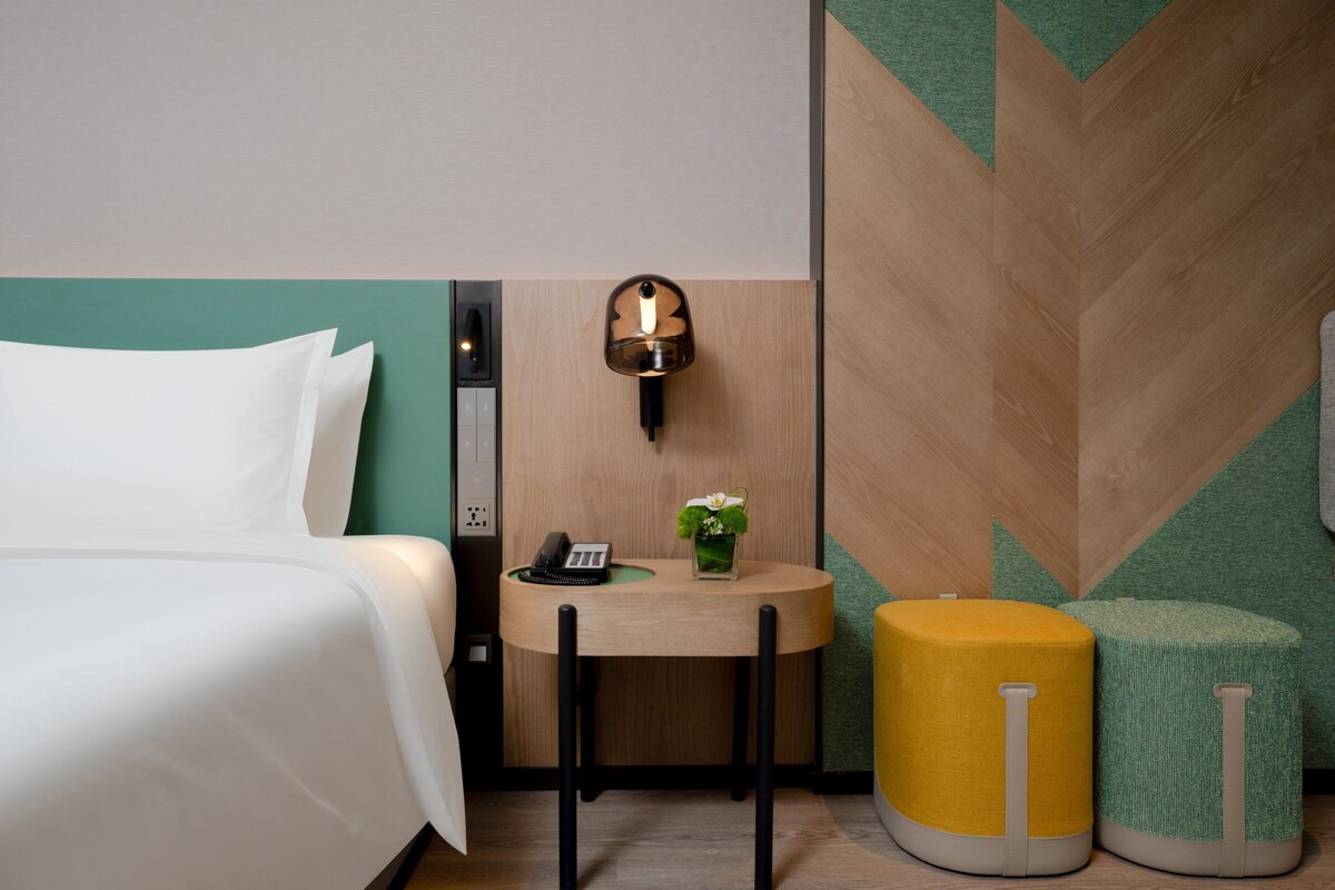 Una habitación de hotel con cama, una mesita con teléfono y un florero, y dos otomanas.
