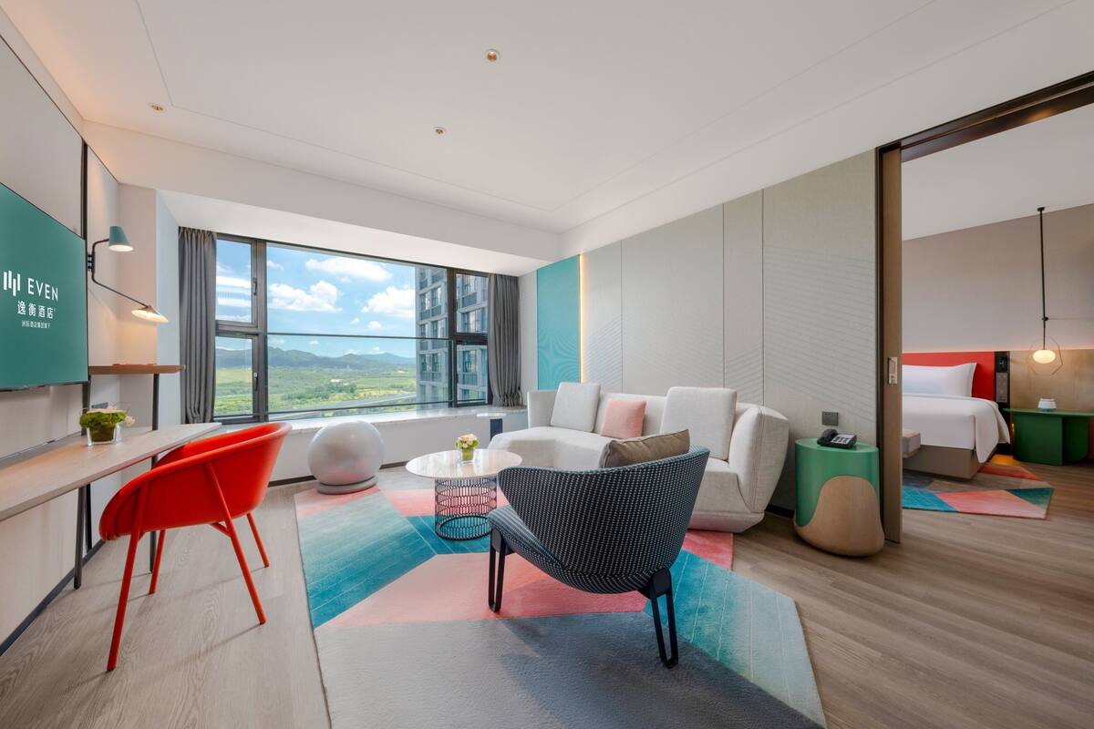 A modern hotel room with a large window, a sofa, a dining area, and a bed.