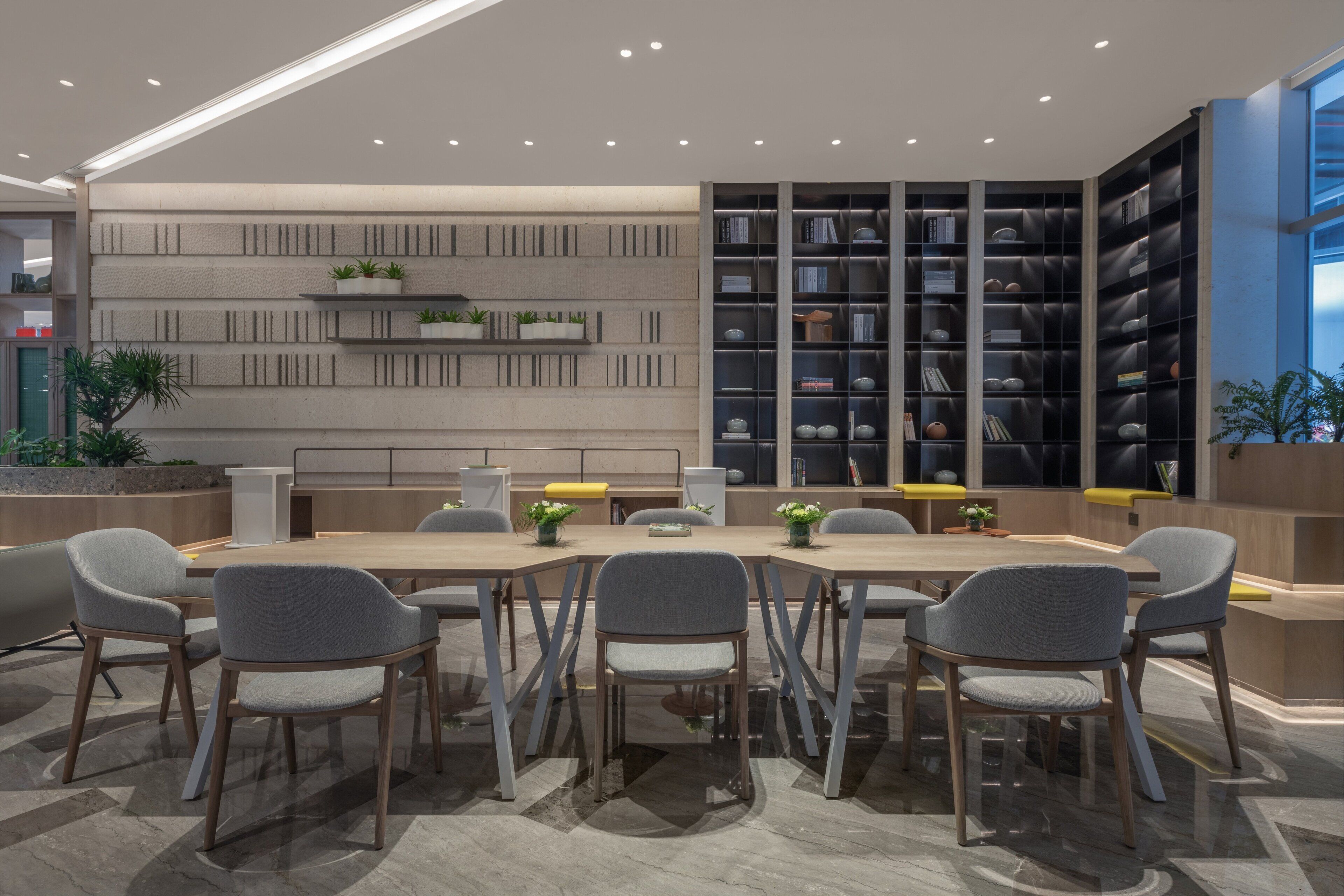 Una sala de conferencias moderna con una larga mesa, sillas grises y una estantería con libros y objetos decorativos.