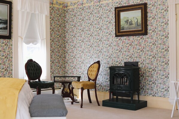 A room with floral wallpaper, a wooden chair, a table, a bed, and a vintage stove. - The Adams Pragge House (Port Townsend)
