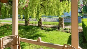 A covered patio area with wicker furniture, a view of a well-maintained garden, and a metal fence in the background. - The Adams Pragge House (Port Townsend)
