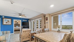 Amplia sala de estar con una mesa de comedor, sillas y un ventilador de techo. Hay un televisor sobre un mueble bajo y un ventanal con vistas al exterior.