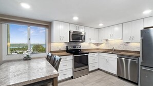 Una cocina moderna con gabinetes blancos, electrodomésticos de acero inoxidable, encimera de granito y vistas al exterior.
