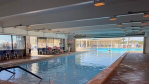 Una piscina interior con agua cristalina, suelo de baldosas y una zona junto a la piscina con sillas y sombrillas.