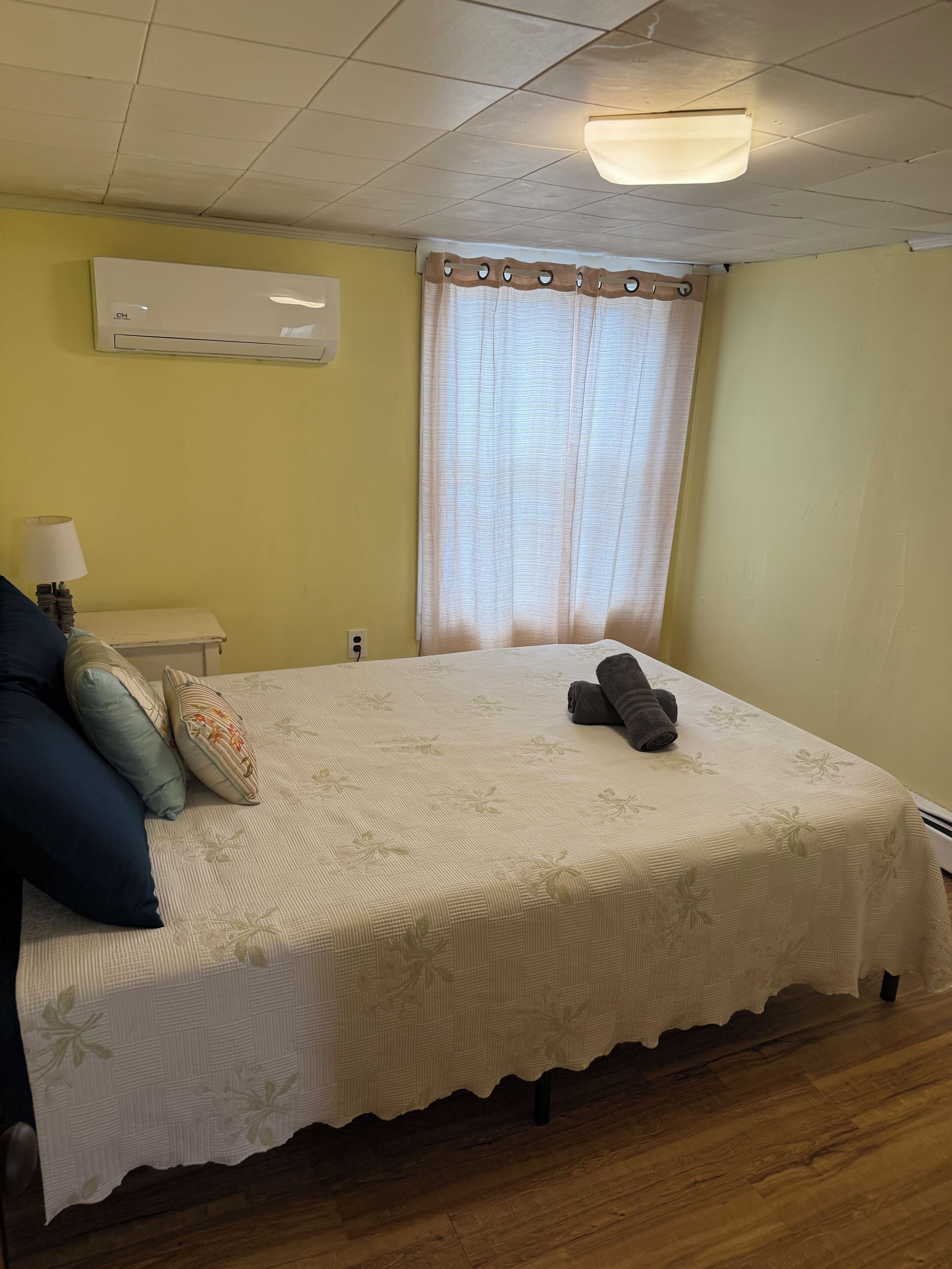 A bedroom with a bed, a nightstand, a lamp, a window with curtains, and a ceiling light.