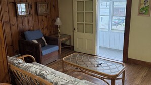 Ein Zimmer mit Holzboden, einem Glastisch, einem Korbssessel, einem blauen Sessel und einer zur Außenseite führenden Tür.