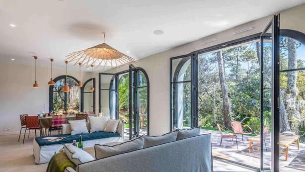 A spacious living area with a large sofa, dining table, and chairs, large windows offering a view of the outdoors.