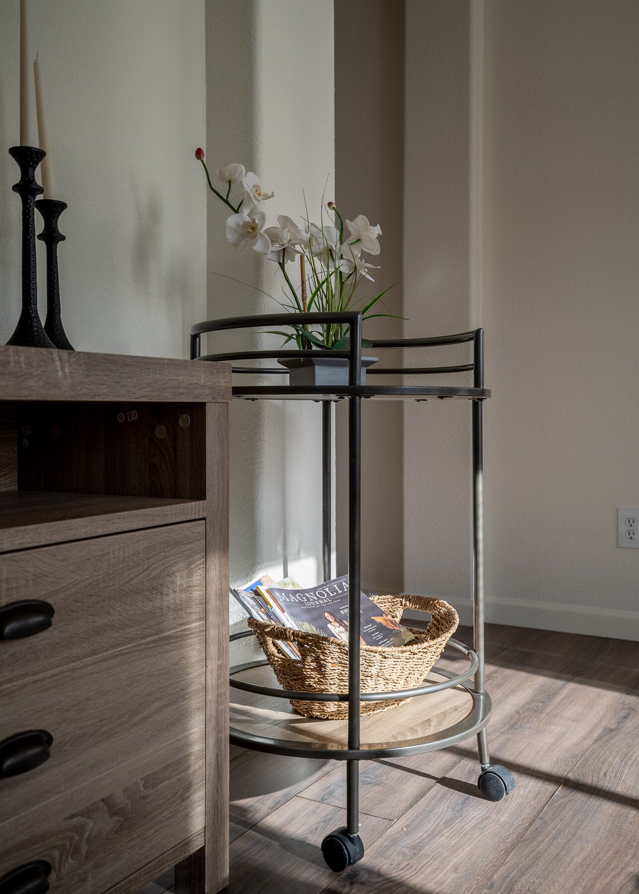 A wooden dresser with a metal cart holding a basket and a vase with white flowers.